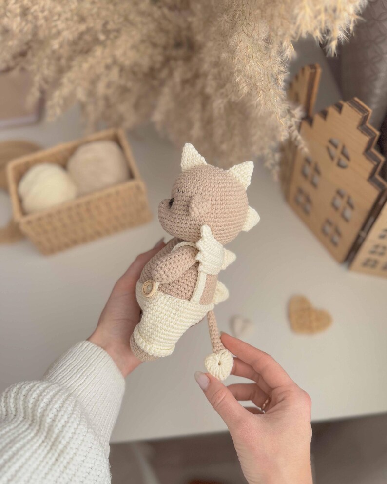 May include: A crocheted plush toy in the shape of a brown and white dragon wearing a white crocheted overall. The dragon has a long tail and a small, pointed snout.