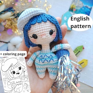 May include: A small crocheted doll with blue hair, a light blue beret, and a striped outfit featuring a wave pattern. The doll holds a silver metallic tassel. Included is a black and white colouring page with the text English pattern + colouring page.