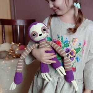 May include: A young girl holds a crocheted purple and brown sloth toy. The sloth has a white face with black eyes and a brown nose.