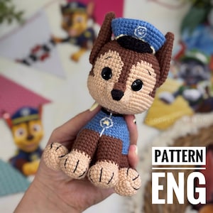 May include: A handmade crochet toy of a cartoon police dog character, featuring brown, tan, and blue colors. The dog wears a blue police hat and uniform. The text "PATTERN ENG" is visible in the image.