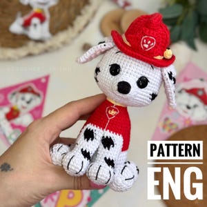 May include: Crocheted white and black spotted puppy wearing a red firefighter hat and uniform. The puppy has black eyes and a black nose. The image includes the text "PATTERN ENG".