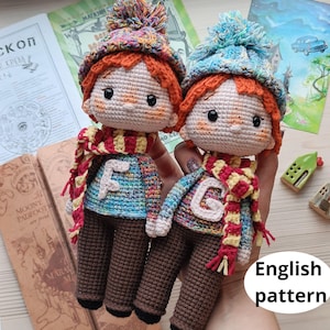 Może przedstawiać: Dwie szydełkowe lalki z czerwonymi włosami, ubrani w kolorowe swetry i szaliki z literami "F" i "G". Lalki mają brązowe spodnie i czarne oczy. Lalki trzymają się za ręce i stoją na stole. Wzór w języku angielskim.