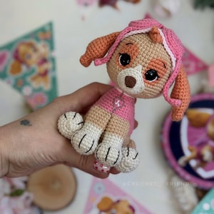 Puede incluir: Un peluche de ganchillo de Skye de Paw Patrol. El juguete es principalmente rosa, beige y blanco, con ojos negros y una nariz marr&oacute;n. El juguete est&aacute; sentado, con sus patas visibles. El fondo incluye pancartas decorativas con personajes de Paw Patrol.
