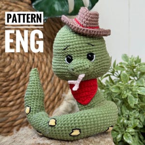 Op de afbeelding: Gehaakte groene slang met een bruine cowboyhoed en een rode bandana. De slang heeft zwarte ogen en een roze tong die uitsteekt. De slang zit op een geweven oppervlak.