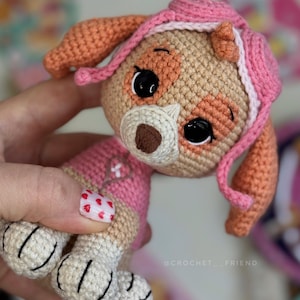 Puede incluir: Un peluche de Skye de Paw Patrol hecho a mano con ganchillo. El juguete presenta una cabeza de color canela con detalles en rosa, ojos negros grandes y una nariz marr&oacute;n. El peluche lleva un atuendo rosa y tiene patas blancas. El juguete se sostiene en una mano.