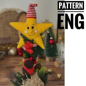 May include: A handmade Christmas tree topper featuring a smiling yellow star with a red and white striped hat. The star is crocheted and sits atop a small tree with red accents. The text "PATTERN ENG" is visible.