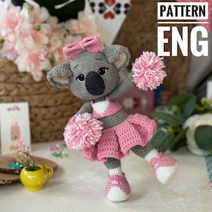 May include: A crocheted koala wearing a pink cheerleader outfit with pom-poms. The koala has a grey body, black eyes, and a pink bow. The outfit has a white heart on the chest and a pink skirt. The koala is standing on a white surface. PATTERN ENG