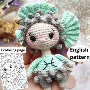 Könnte beinhalten: Eine gehäkelte Puppe mit einem hellen mintgrünen und grauen strukturierten Outfit, passender Kopfbedeckung, großen schwarzen Augen und rosa Wangen. Die Puppe hat ein schwarzes Fische-Symbol auf der Brust. Enthält eine schwarz-weiße Malvorlage.