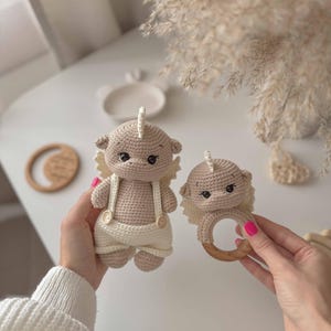 May include: Two crocheted plush toys in the shape of a baby animal with a wooden ring rattle. The larger toy is wearing overalls and the smaller toy is attached to the wooden ring.