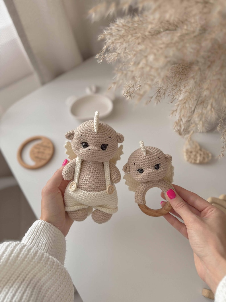 May include: Two crocheted plush toys in the shape of a baby animal with a wooden ring rattle. The larger toy is wearing overalls and the smaller toy is attached to the wooden ring.