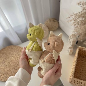 May include: Two crocheted stuffed toys in the shape of dragons wearing white overalls with buttons. One dragon is green and the other is brown.