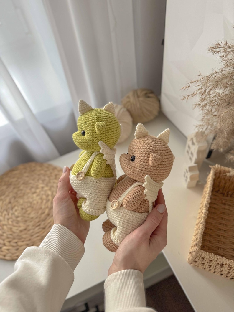 May include: Two crocheted stuffed toys in the shape of dragons wearing white overalls with buttons. One dragon is green and the other is brown.