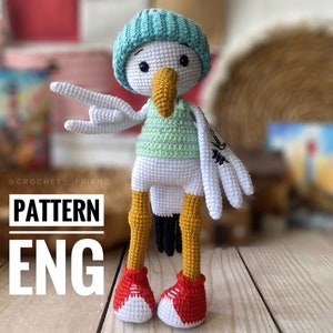 Könnte beinhalten: Eine gehäkelte Amigurumi-Möwe, die ein blau-weiß gestreiftes Hemd, einen blauen Hut und rote Schuhe trägt. Die Möwe hält ihre Flügel ausgestreckt und hat einen freundlichen Ausdruck. Der Text "PATTERN ENG" ist auf dem Bild sichtbar.