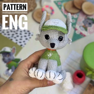 Puede incluir: Juguete de peluche de ganchillo gris y verde de un personaje de dibujos animados, con el texto "PATTERN ENG" en la esquina superior izquierda. El juguete tiene grandes ojos negros, un sombrero verde y blanco y una camisa verde. El juguete se sostiene en una mano.