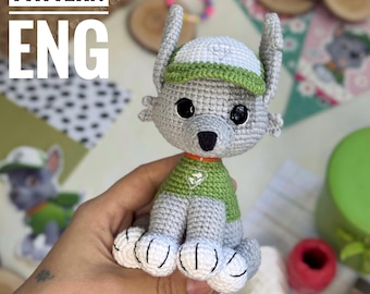 Può includere: Peluche all'uncinetto grigio e verde di un personaggio dei cartoni animati, con la scritta "PATTERN ENG" nell'angolo in alto a sinistra. Il giocattolo ha grandi occhi neri, un cappello verde e bianco e una camicia verde. Il giocattolo è tenuto in mano.