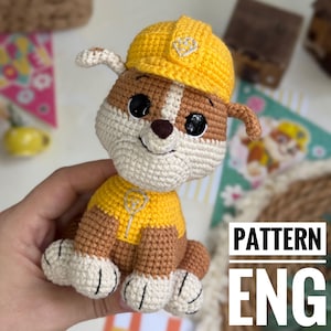 Op de afbeelding: Gehaakte speelgoedhond in geel en bruin, met een gele helm en shirt. De hond heeft zwarte ogen en een wit gezicht en poten. De tekst "PATTERN ENG" wordt in het wit op een zwarte achtergrond weergegeven.