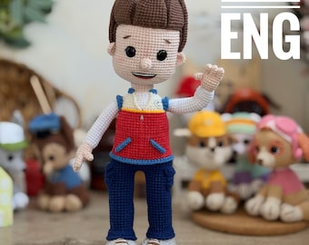 Può includere: Bambola all'uncinetto di un personaggio dei cartoni animati, con un gilet rosso, pantaloni blu e scarpe bianche. La bambola ha i capelli castani e sta salutando. Il testo "PATTERN ENG" è visibile. Altre bambole simili sono sullo sfondo.