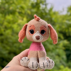 Puede incluir: Un juguete de perro cachorro de ganchillo en color beige, rosa y blanco. El juguete hecho a mano tiene ojos negros grandes, una nariz marr&oacute;n y orejas rosas. El perro lleva una camisa rosa y est&aacute; sentado. El fondo es verde borroso.