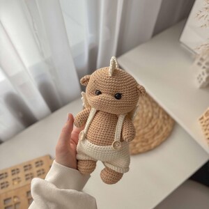 May include: A crocheted stuffed animal in the shape of a bear wearing a white crocheted outfit with suspenders and buttons. The bear has brown fur and a white horn on its head.