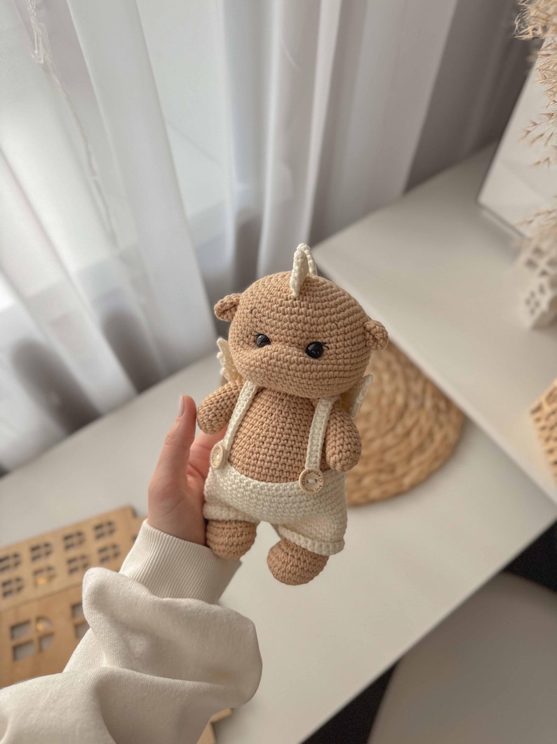 May include: A crocheted stuffed animal in the shape of a bear wearing a white crocheted outfit with suspenders and buttons. The bear has brown fur and a white horn on its head.