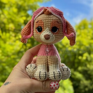 Puede incluir: Un peluche de Skye de Paw Patrol hecho a mano con ganchillo. El juguete presenta una combinaci&oacute;n de colores rosa y beige, con ojos negros grandes y una peque&ntilde;a nariz marr&oacute;n. El juguete se sostiene frente a un fondo verde borroso.