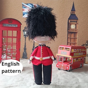 May include: A handmade crochet doll dressed as a British Royal Guard, wearing a red jacket with white trim and black pants. The doll has a large black hat. The background includes a red telephone booth, a double-decker bus, and a Big Ben replica.