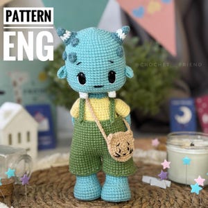 May include: A crocheted blue monster doll with yellow and green clothing and a brown purse. The doll has a friendly expression and is standing on a woven surface.