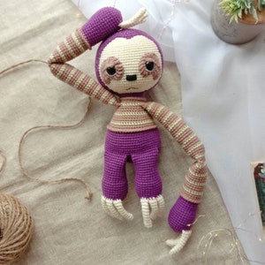 May include: A purple and brown crocheted sloth toy with a striped pattern. The sloth has a long, curled tail and large, black eyes.
