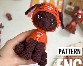 May include: Crocheted dog toy in orange and brown. The dog is wearing an orange outfit with a white emblem and a matching hood. The toy is handmade and has large black eyes. The text "PATTERN ENG" is visible in the image.