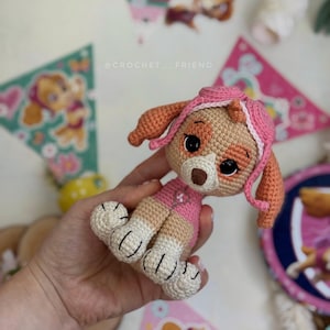 Puede incluir: Mu&ntilde;eca Skye de Paw Patrol hecha a ganchillo. La mu&ntilde;eca es beige, rosa y naranja, con ojos negros y nariz marr&oacute;n. Lleva un atuendo y sombrero rosa. La mu&ntilde;eca est&aacute; sujeta en una mano. Hay banderines decorativos.