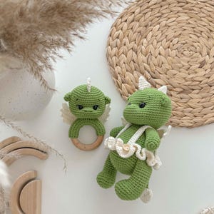 May include: Two green crocheted dragon toys. One is a rattle with a wooden ring and the other is a plush toy with a white crocheted skirt and a button.