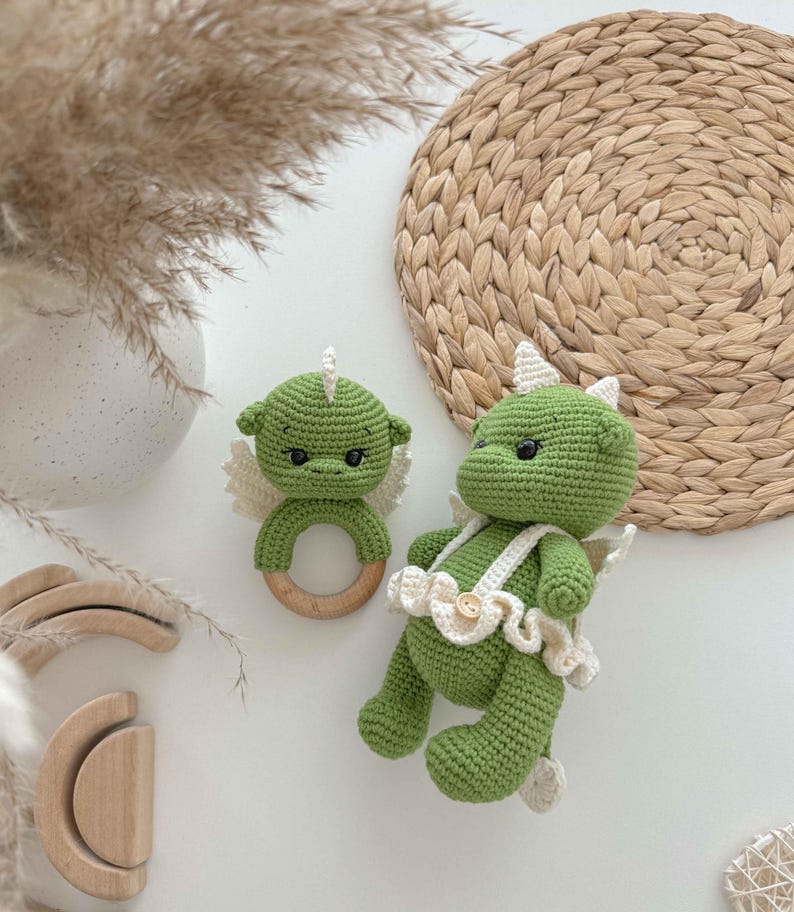 May include: Two green crocheted dragon toys. One is a rattle with a wooden ring and the other is a plush toy with a white crocheted skirt and a button.