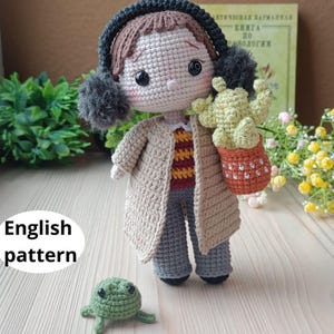 May include: A handmade crochet doll wearing a tan coat, gray pants, and a striped shirt. The doll has brown hair, black earmuffs, and is holding a cactus in a terracotta pot. A small green turtle is at the doll's feet. The text "English pattern" is visible.