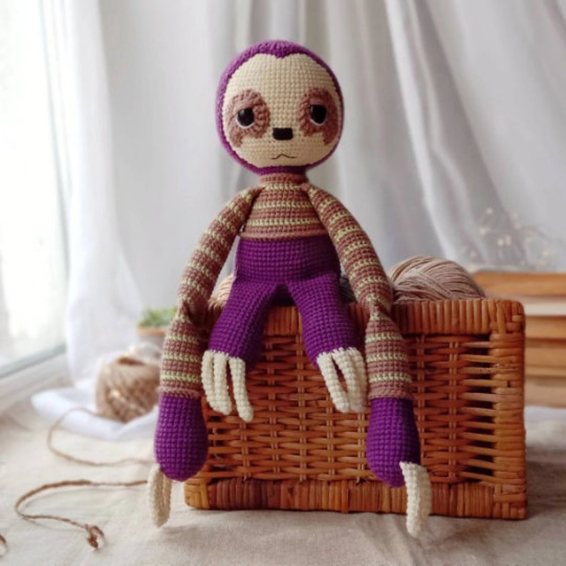 May include: A crocheted sloth toy with purple and brown stripes, sitting on a wicker basket. The sloth has a beige face with brown eyes and a small nose.
