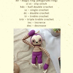 May include: Purple and brown crocheted sloth amigurumi toy. The toy is sitting on a beige fabric surface. The toy has a sleepy expression and is holding its arm up to its head. The toy is made with a single crochet stitch and has a striped pattern on its arms and torso.