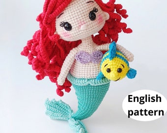 Puede incluir: Muñeca de sirena de ganchillo hecha a mano, con cabello rojo, top morado y cola turquesa. La muñeca sostiene un pez amarillo y azul. El texto "English pattern" es visible.