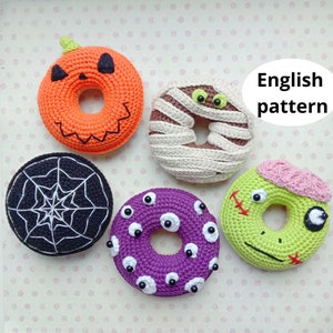 Può includere: Cinque ciambelle all'uncinetto a tema Halloween. Le ciambelle sono arancioni, marroni, nere, viola e verdi. Le ciambelle sono decorate con una faccia di zucca, bende da mummia, una ragnatela, occhietti e una faccia di zombie.