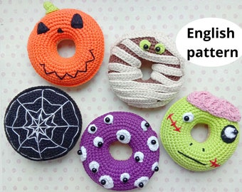 May include: Five crocheted Halloween-themed doughnuts. The doughnuts are orange, brown, black, purple, and green. The doughnuts are decorated with a pumpkin face, mummy bandages, a spiderweb, googly eyes, and a zombie face.