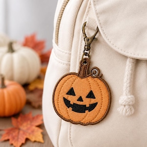 May include: An orange fabric pumpkin keychain with black embroidered jack-o'-lantern facial features, including triangular eyes and a jagged mouth, attached to a cream-colored backpack with a bronze-toned metal lobster clasp.