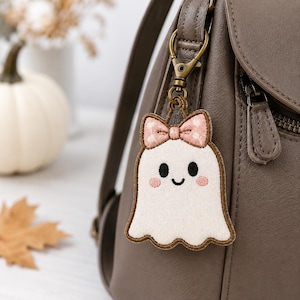 May include: A white ghost-shaped fabric keychain featuring a pink polka-dot bow, black embroidered eyes, a small smile, and pink cheeks. The ghost is attached to a bronze-colored metal lobster clasp hook, which is clipped onto a brown leather backpack.