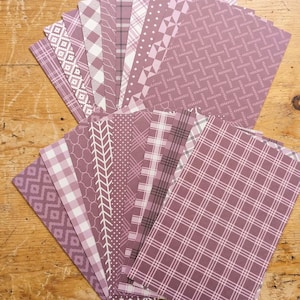 May include: A collection of patterned paper sheets in shades of mauve and white. The designs include geometric shapes, plaid, and chevron patterns. The papers are arranged on a wooden surface, showcasing the variety of designs.