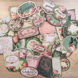 May include: A collection of vintage-style paper ephemera with floral and botanical designs. The pieces include labels, tags, and circular elements, featuring roses and text like "Journal," "Garden," and "In Bloom."