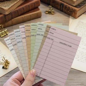 May include: A hand holds a fan of vintage-style library cards in various pastel colors. Each card has lines for "Author," "Title," and "Date Due." The cards are arranged in a fanned-out pattern, with the text "Borrower's Name" visible on one.