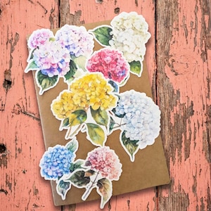 May include: A collection of colorful hydrangea flower stickers in shades of pink, purple, yellow, white, and blue, with green leaves. The stickers are arranged on a brown notebook against a weathered, pink wooden background.