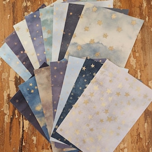 May include: A collection of celestial-themed stationery. Each sheet features a watercolor background in shades of blue, purple, and gray, adorned with gold star patterns. The papers are arranged in a fan shape, showcasing the variety of designs.