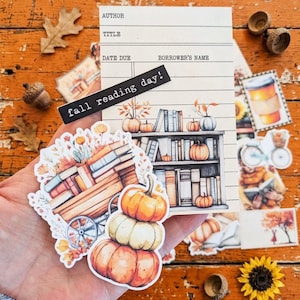 May include: A collection of paper stickers featuring a library checkout card with the text fall reading day, a wooden cart filled with books and autumn flowers, and a stack of three pumpkins in orange and cream colors with fall foliage.