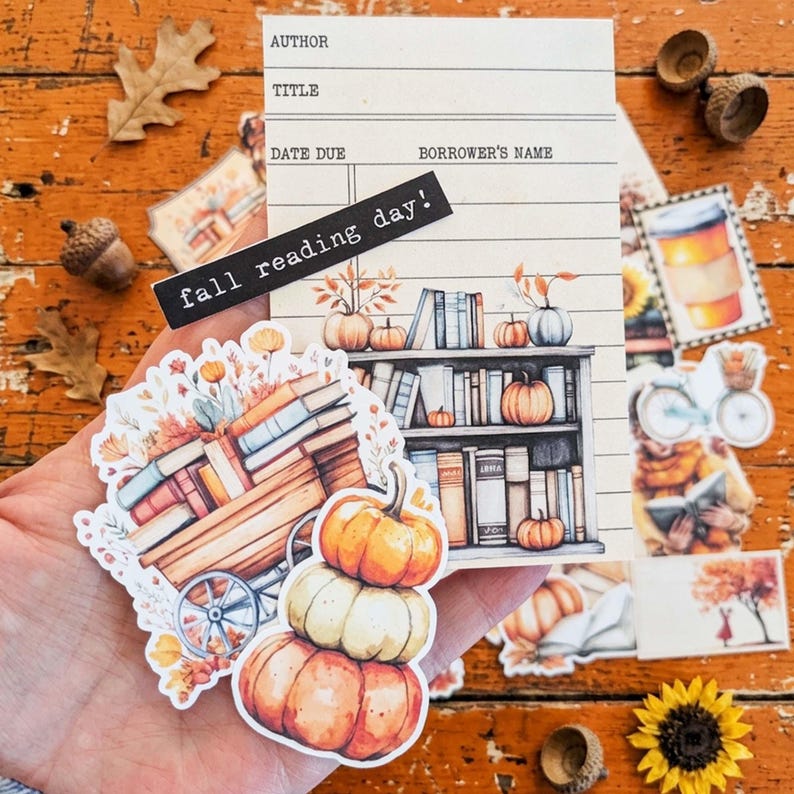 May include: A collection of paper stickers featuring a library checkout card with the text fall reading day, a wooden cart filled with books and autumn flowers, and a stack of three pumpkins in orange and cream colors with fall foliage.