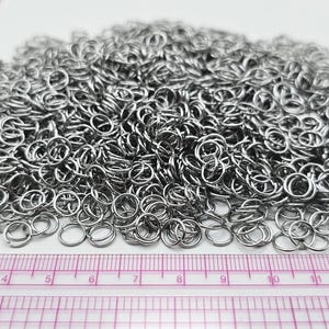 May include: A large pile of small, circular, silver-coloured metal jump rings. A clear plastic ruler is positioned along the bottom and left side of the pile, showing the rings measure approximately 0.6 cm in diameter for individual units.