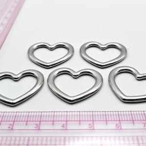May include: Five silver-colored heart-shaped charms are arranged on a white surface. A pink ruler with centimeter and inch markings is in the foreground. The charms are made of a shiny metal.