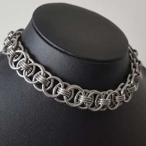 Puede incluir: Un collar de cota de malla plateado exhibido en un maniquí negro. El collar está hecho de anillos entrelazados, creando una apariencia gruesa y texturizada. El collar, estilo gargantilla, está diseñado para quedar cerca del cuello. Mide aproximadamente 40 cm.